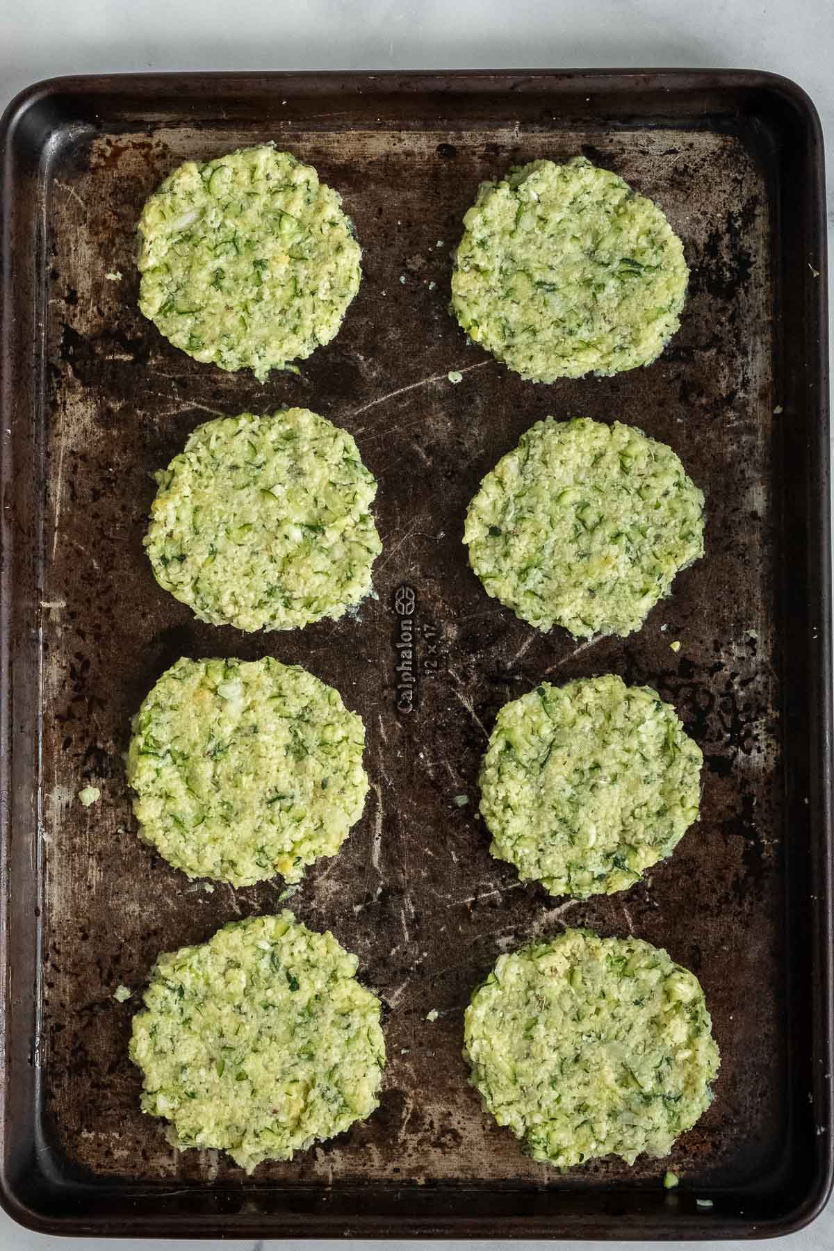 eight uncooked zucchini fritters on a baking sheet