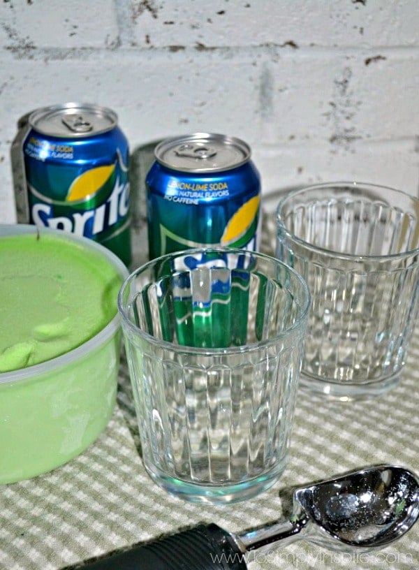 Two glasses on a green checked placemat with a bowl of lime sherbet and two cans of sprite.