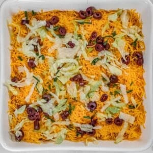 A delightful Layered Taco Dip featuring shredded cheddar cheese, chopped lettuce, sliced olives, and green onions.