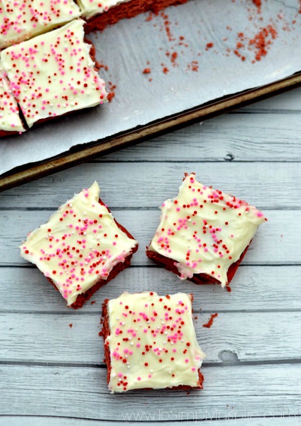 Three red velvet cake cookie bars topped with cream cheese frosting and sprinkles on a grey wood table