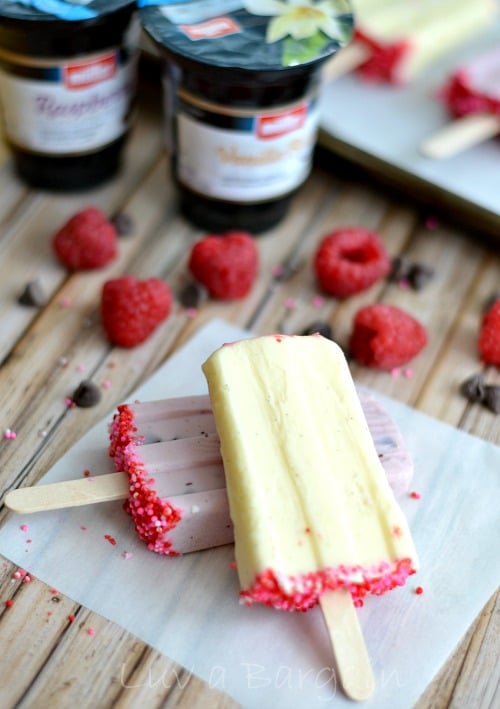 yellow and pink yogurt popsicles on a piece of parchment paper with fresh raspberries laying beside