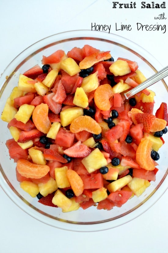 This Fruit Salad with Honey Lime Dressing is a deliciously refreshing addition to any meal. It's the best for getting more fruit into everyone's day and perfect for any Summer BBQ. a big glass bowl filled with a mixture of oranges, watermelon, pineapple, strawberries and blueberries