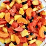 A bowl of oranges, watermelon, pineapple, strawberries and blueberries in a glass bowl with a spoon