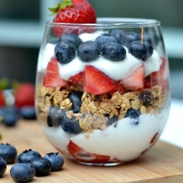 A glass layered with yogurt, blueberries, granola and strawberries on a wooden cutting board