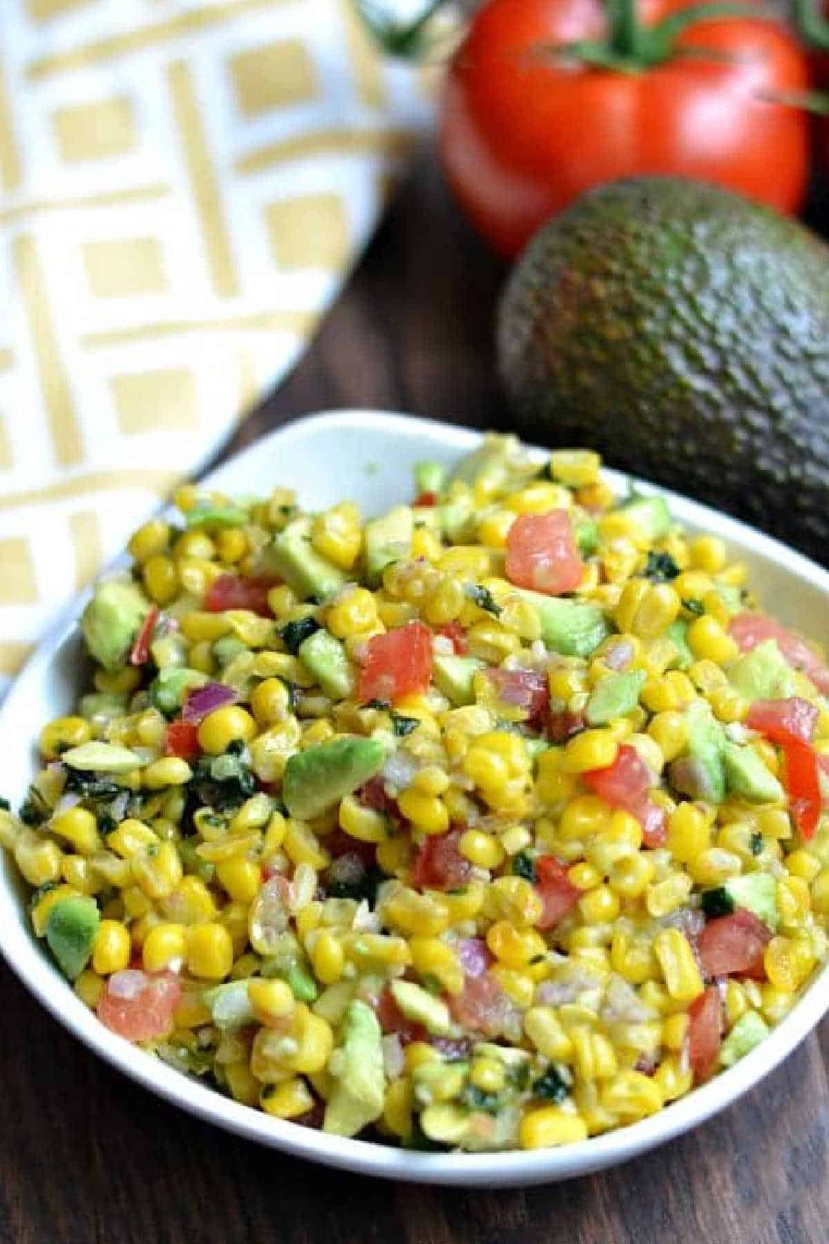 Corn Salsa recipe in a white bowl with an avocado and tomato beside it.