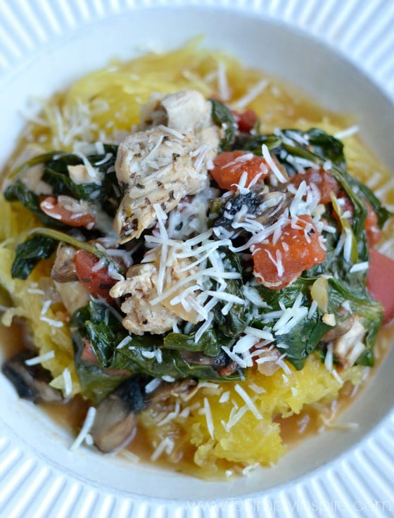 a white bowl with cooked spaghetti squash topped with cooked spinach, tomatoes, chicken and parmesan cheese