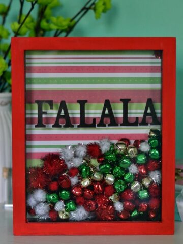 a red shawdow box frame filled with red green and white bells and pom moms that say FA LA LA in the front of the glass