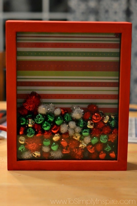 a shadowbox frame with a green and red striped background and filled with green red and white pom moms and bells 