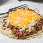A plate of spaghetti topped with Cincinnati Chili Recipe ground beef sauce, shredded cheese, and chopped onions, with a fork placed on the side.