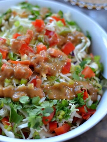 A close up of a bowl of lettuce with diced red peppers and a Thai peanut butter dressing drizzled on top