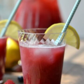 A close up of a cup of raspberry blueberry lemonade with a light blue straw and a lemon wedge