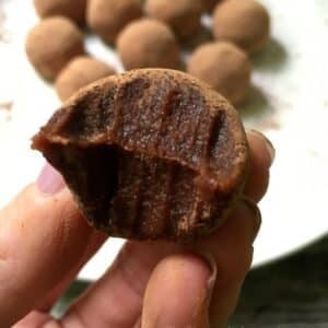 A hand holds a chocolate date truffle with a bite taken out, revealing its rich, dense interior. More truffles are visible on a plate in the background.