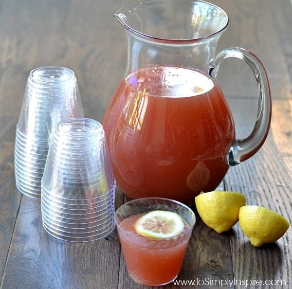 A glass pitcher of red punch with 2 lemon halves and a stack of plastic cups