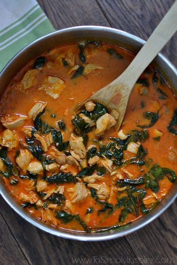 A pot of soup and a spoon with chicken and chickpeas in a red sauce