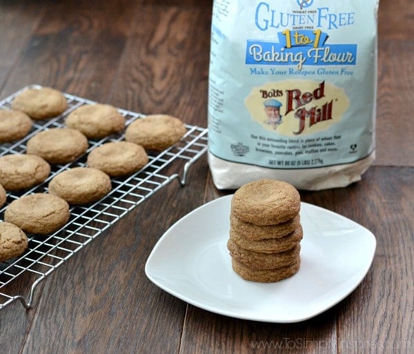 A stack of 5 molasses cookies on a white plate with a cooling rack of cookies beside and a bog of bobs red mill gluten free flour. 