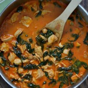 A pot of hearty curry chicken stew with chickpeas and leafy greens, served with a wooden spoon.