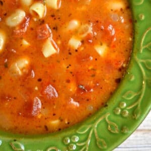 Close-up of a bowl of pasta fagioli soup with herbs, diced tomatoes, small pasta pieces, and pepperoni, served in a green bowl.