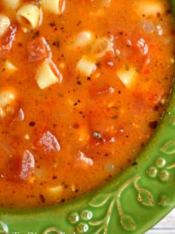 Close-up of a bowl of pasta fagioli soup with herbs, diced tomatoes, small pasta pieces, and pepperoni, served in a green bowl.