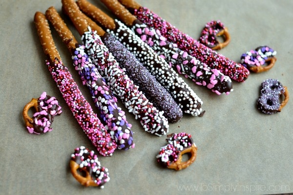 Seven Chocolate Covered Pretzel rods covered in valentines sprinkles laying on parchment paper with pretzels knots beside