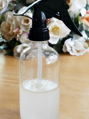 Glass Spray bottle with black top half filled with liquid homemade bug spray with flowers in the background