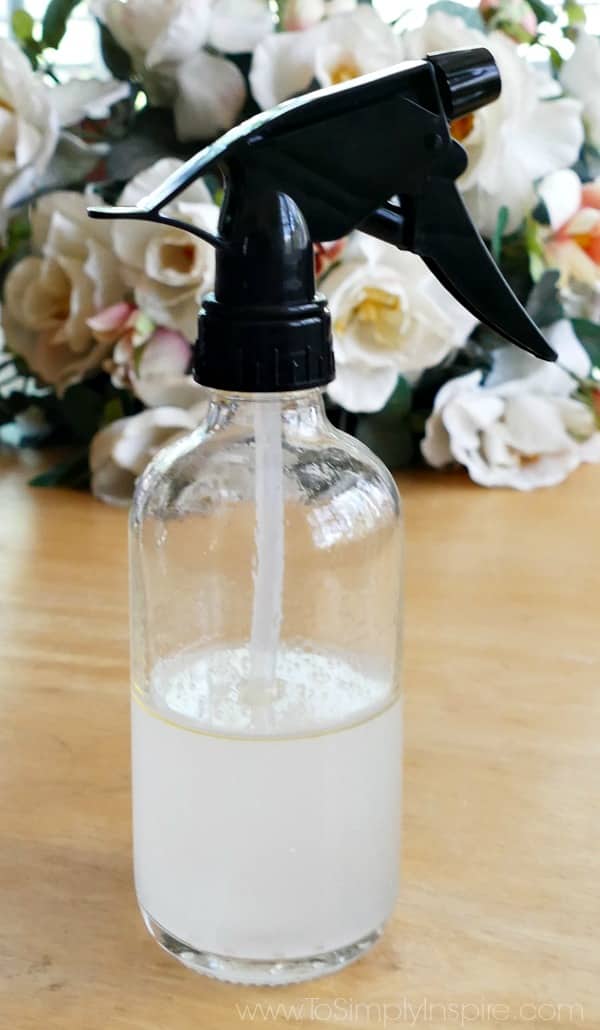 A close up of a Glass Spray bottle with black top half filled with liquid homemade bug spray with flowers in the background