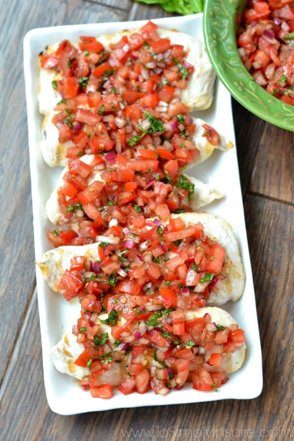 closeup of 5 chicken breasts topped with bruschetta tomato sauce on a white rectangle plate