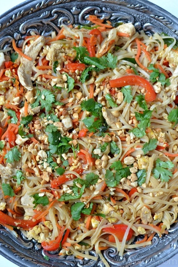 A dish is filled rice noodles, red pepper strips, cilantro, chicken and peanuts