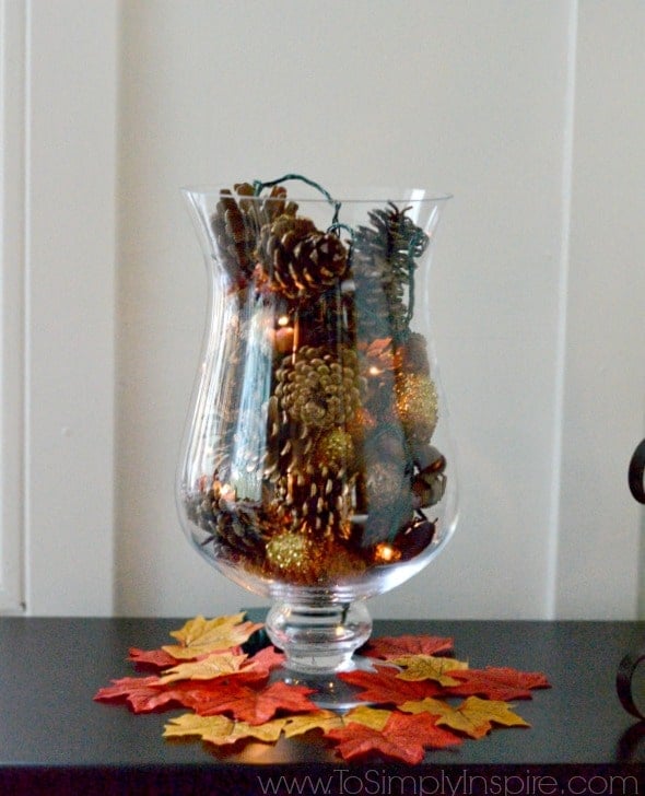 A glass vase on a table filled with pinecones and fairy lights