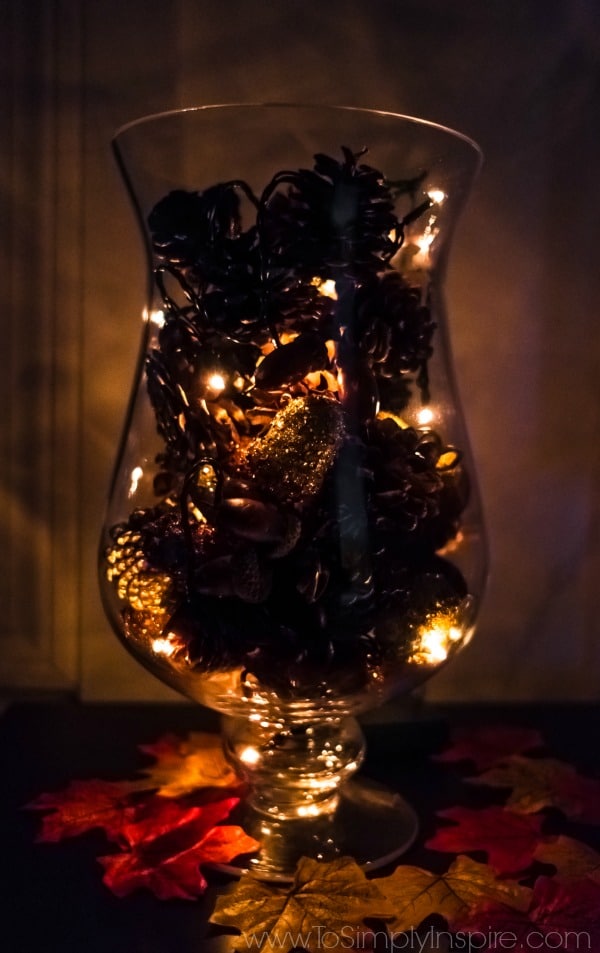 A glass vase on a table filled with pinecones and fairy lights turned on 