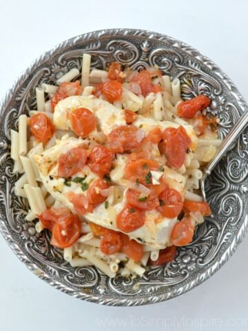 A silver bowl with cod fish topped with cherry tomatoes and basil on top of penne pasta