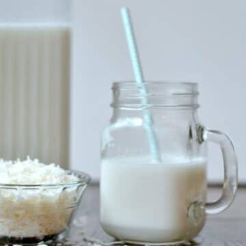 A glass with a handle of Coconut Milk with a blue straw, a small glass bowl of shredded coconut and a glass pitcher of milk