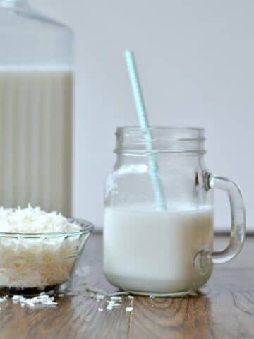 A glass with a handle of Coconut Milk with a blue straw, a small glass bowl of shredded coconut and a glass pitcher of milk