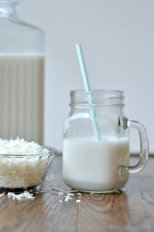 A glass with a handle of Coconut Milk with a blue straw, a small glass bowl of shredded coconut and a glass pitcher of milk 