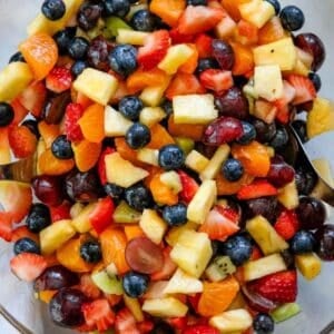 A bowl of Fruit Salad with Honey Lime Dressing featuring fresh blueberries, strawberries, grapes, kiwi, pineapple, and mandarin orange segments, with serving utensils visible.