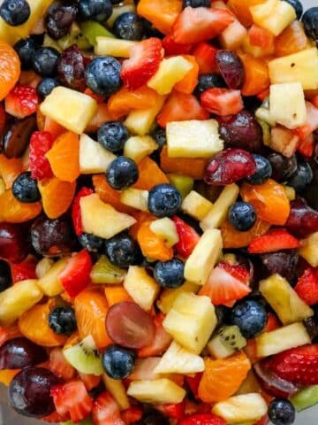 A bowl of Fruit Salad with Honey Lime Dressing featuring fresh blueberries, strawberries, grapes, kiwi, pineapple, and mandarin orange segments, with serving utensils visible.
