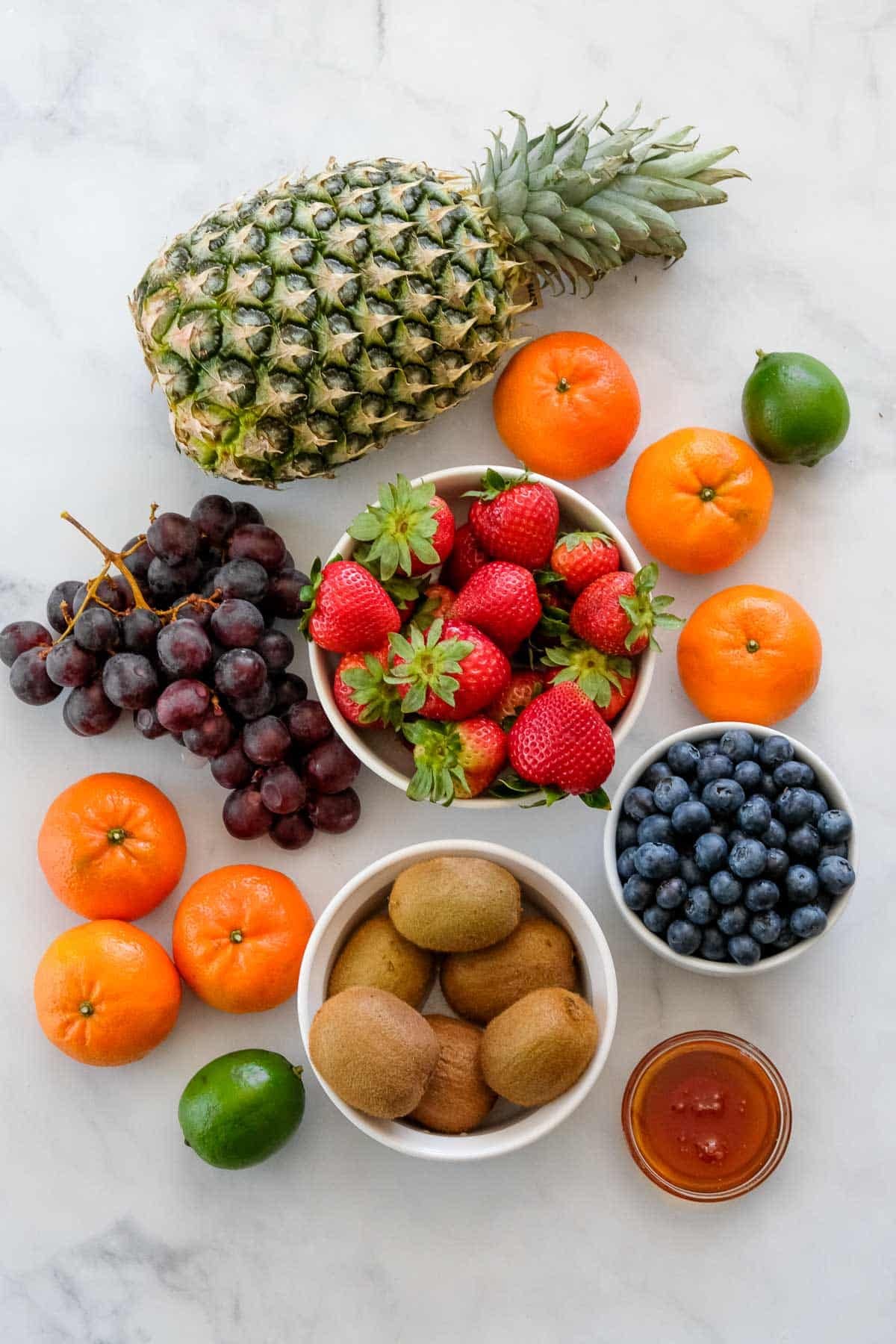 A variety of fresh fruits including pineapple, strawberries, grapes, blueberries, kiwis, mandarins, and limes are beautifully arranged with a small bowl of honey for a vibrant Fruit Salad with Honey Lime Dressing on a white surface.