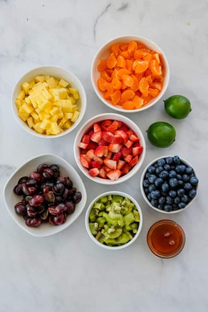 Five bowls with chopped pineapple, mandarin oranges, strawberries, cherries, and kiwi, plus a bowl of blueberries, two whole limes, and a small container of honey—perfect for making a vibrant Fruit Salad with Honey Lime Dressing.