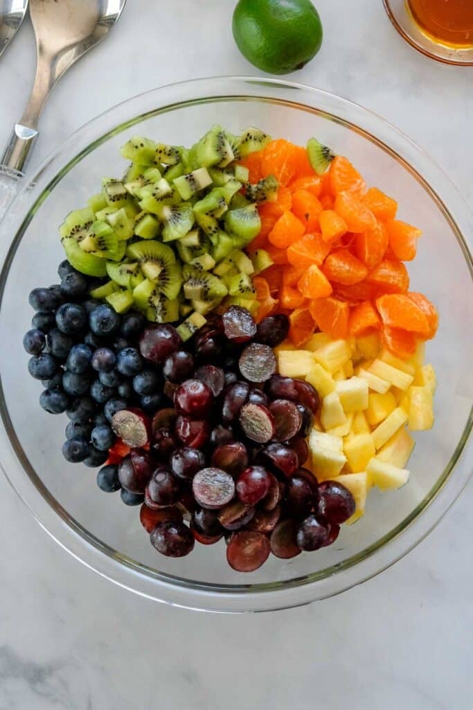 A glass bowl filled with sections of sliced kiwi, mandarin oranges, blueberries, red grapes, and pineapple sits on a marble surface—an inviting Fruit Salad with Honey Lime Dressing. A lime and utensils are nearby.