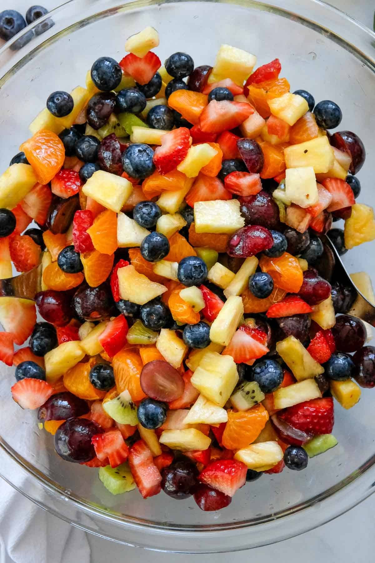 A clear bowl filled with a colorful mix of chopped strawberries, blueberries, grapes, kiwi, pineapple, and mandarin oranges is ready to serve as a refreshing Fruit Salad with Honey Lime Dressing. Serving spoons are included.