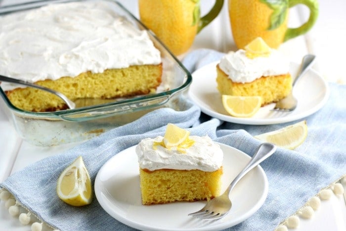 2 slices of lemonade cake with frosting on white plates with whole cake pan and 2 yellow mugs in background