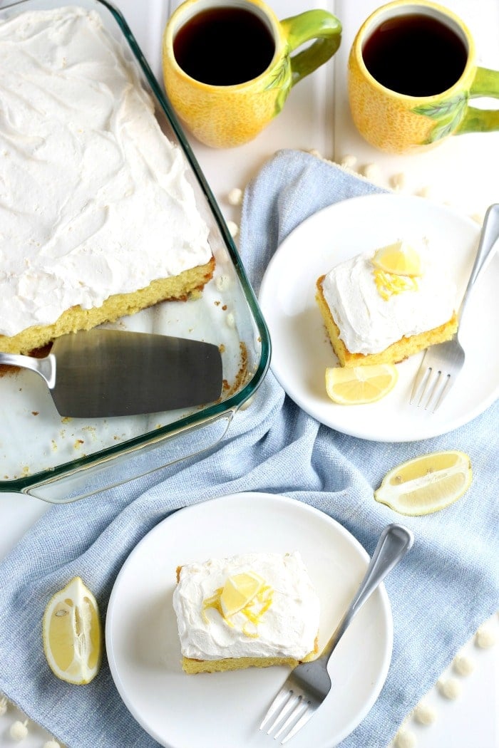 Overhead picture of two slices of lemonade cake with frosting on white plates with cake dish and 2 myellow mugs of coffee in the background