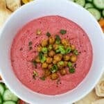 A bowl of pink beet hummus garnished with chickpeas, green onions, and herbs. Surrounded by cucumber slices, bell pepper strips, and pita bread.