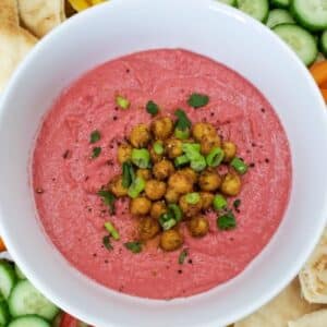 A bowl of pink beet hummus garnished with chickpeas, green onions, and herbs. Surrounded by cucumber slices, bell pepper strips, and pita bread.