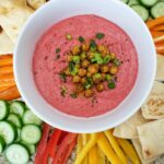 beet hummus in a white bowl surrounded by fresh vegetables