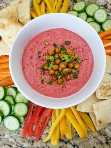 beet hummus in a white bowl surrounded by fresh vegetables