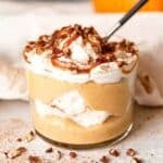 A glass cup filled with layers of pumpkin mousse, whipped cream, and meringue, topped with caramel sauce and chopped pecans, with a spoon inside.
