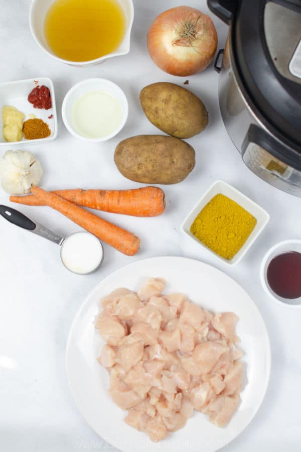 diced raw chicken on a plate with 2 carrots, 2 whole potatoes and measuring cups and spoons on a white counter