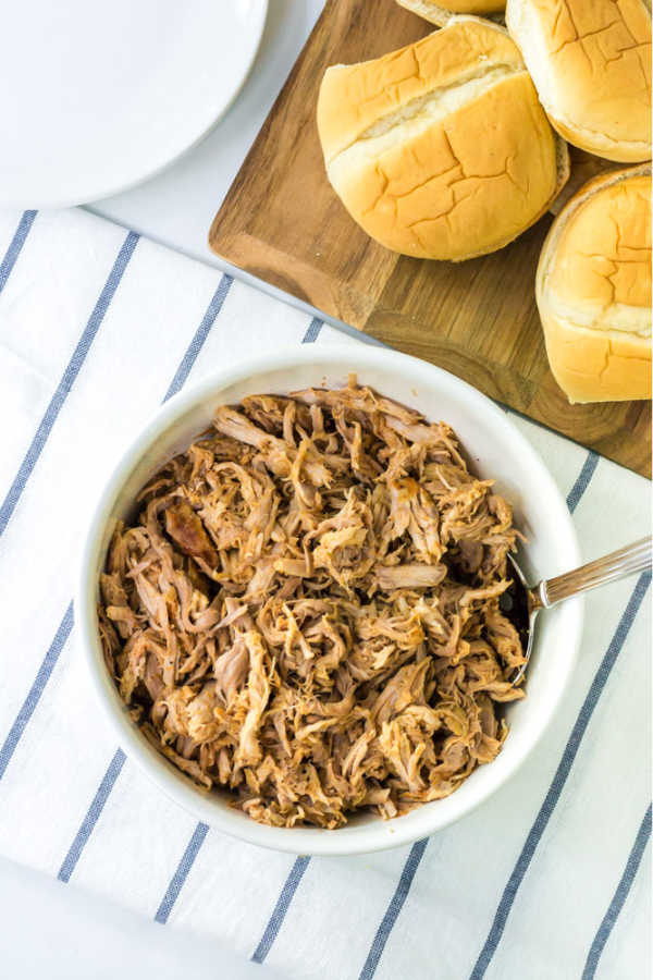 a white bowl full of shredded bbq pulled pork with a stack of buns beside