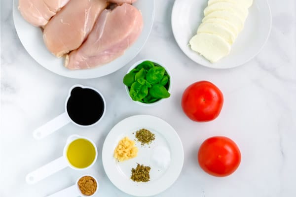 4 raw chicken breasts on a white plate surrounded by fresh mozzarella cheese, fresh basil, 2 tomatoes and spices