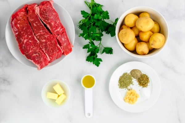 Uncooked steak on a plate, uncooked potatoes in a bowl, fresh parsley, and spices